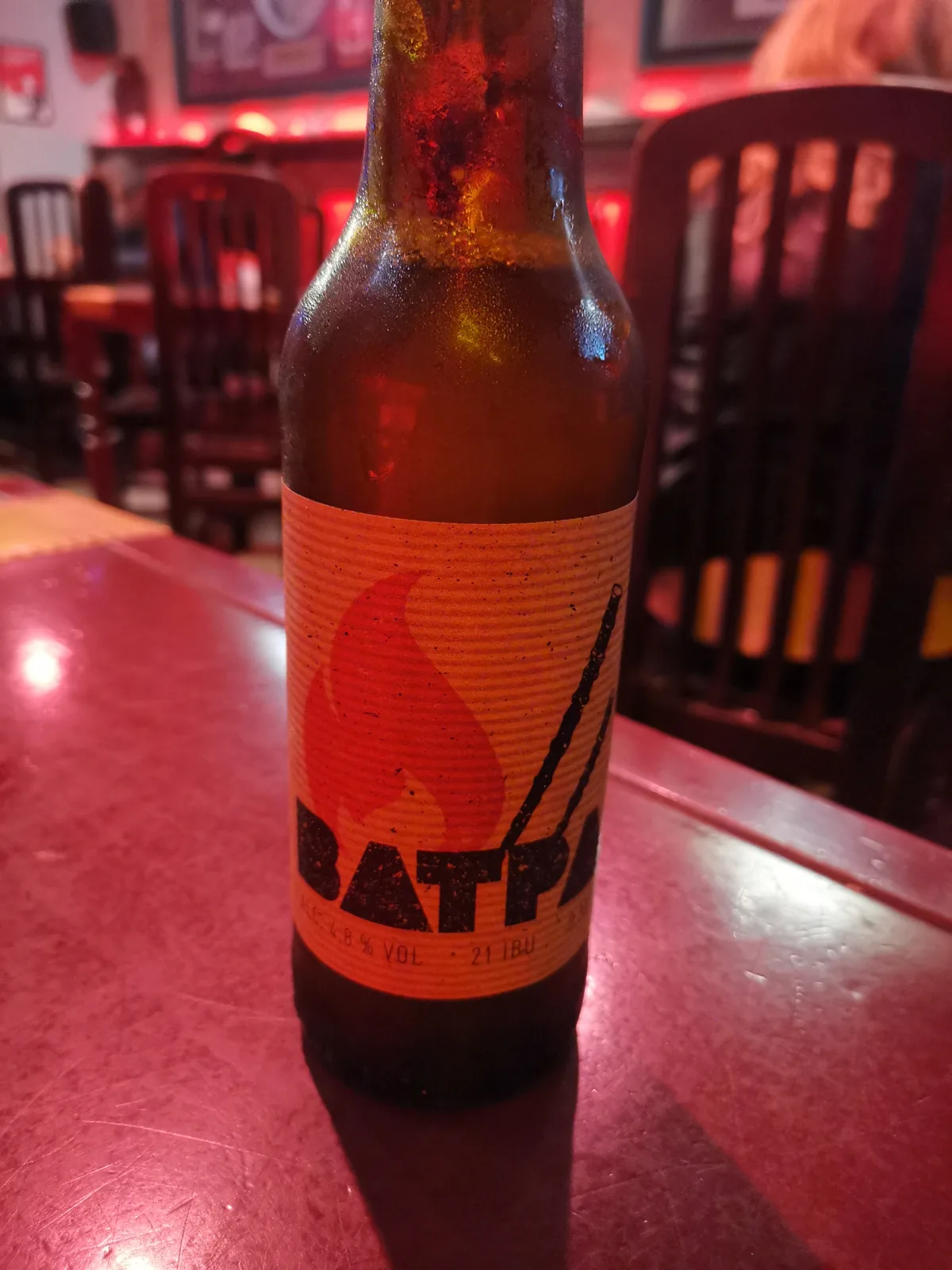 A cerveja Batt apresenta uma graduação alcoólica moderada com amargor de 21.1 IBU. Apesar da etiqueta laranja característica visível na imagem, não há informações suficientemente claras no rótulo para identificar com precisão a cervejaria, o país de origem ou o estilo específico da cerveja. A falta de detalhes legíveis na imagem impede uma análise mais completa das características marcantes dessa bebida.